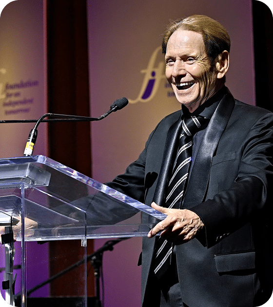 A man with light brown hair in a black tuxedo and striped tie laughs while standing at a clear podium.
