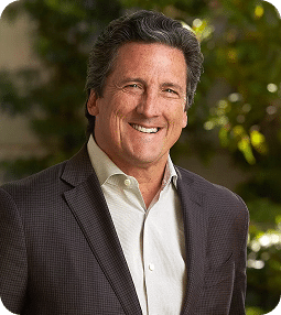 A smiling man with dark, wavy hair wears a gray suit jacket and a light-colored collared shirt.