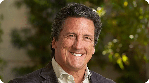 A smiling man with dark, wavy hair and a gray suit jacket looks directly at the viewer, with blurred green foliage behind him.