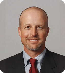 A smiling bald man with a short beard wears a black suit, light blue shirt, and red tie.