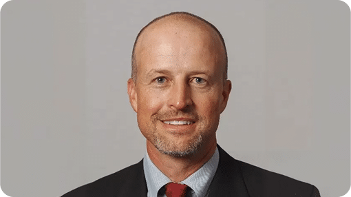 A smiling bald man with a light beard and blue eyes wears a dark suit and a red tie.
