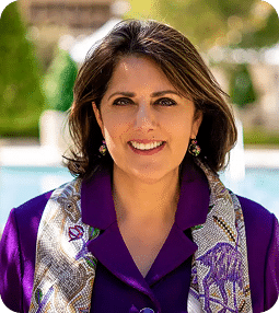 A smiling woman with dark hair wears a purple blazer and a patterned silk scarf outdoors.