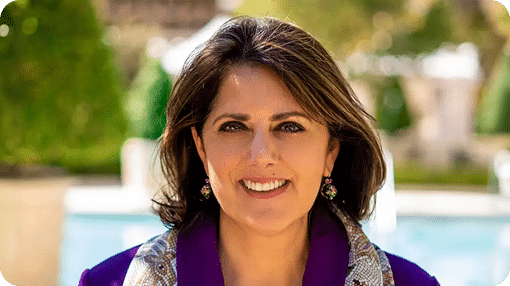 A smiling woman with dark hair and a purple blazer wears colorful earrings, with a blurred garden behind her.