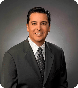 A smiling man with dark hair, wearing a gray suit, patterned tie, and white shirt, against a gray background.