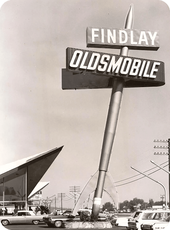 A black and white photo of a rocket-shaped sign for "FINDLAY OLDSMOBILE" towering over a car dealership.