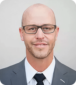 A smiling bald man with a light beard and glasses wears a gray suit and dark tie.