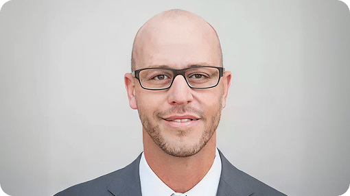 A bald man with a short beard and glasses smiles slightly, wearing a suit jacket and white shirt.