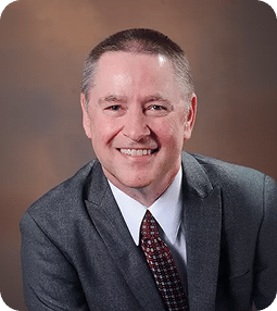 A smiling man with short brown hair wears a gray suit jacket and a patterned red tie.