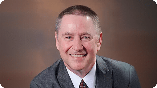 A smiling man with short brown hair, wearing a gray suit, white shirt, and patterned tie.