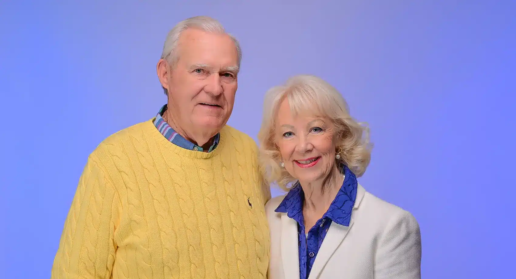 An older man in a yellow cable-knit sweater and a woman in a white blazer smile at the camera.